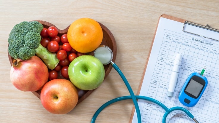 A heart shaped bowl of fruit with a stethoscope, a blood sugar tracker sheet on a clipboard and diabetic equipment